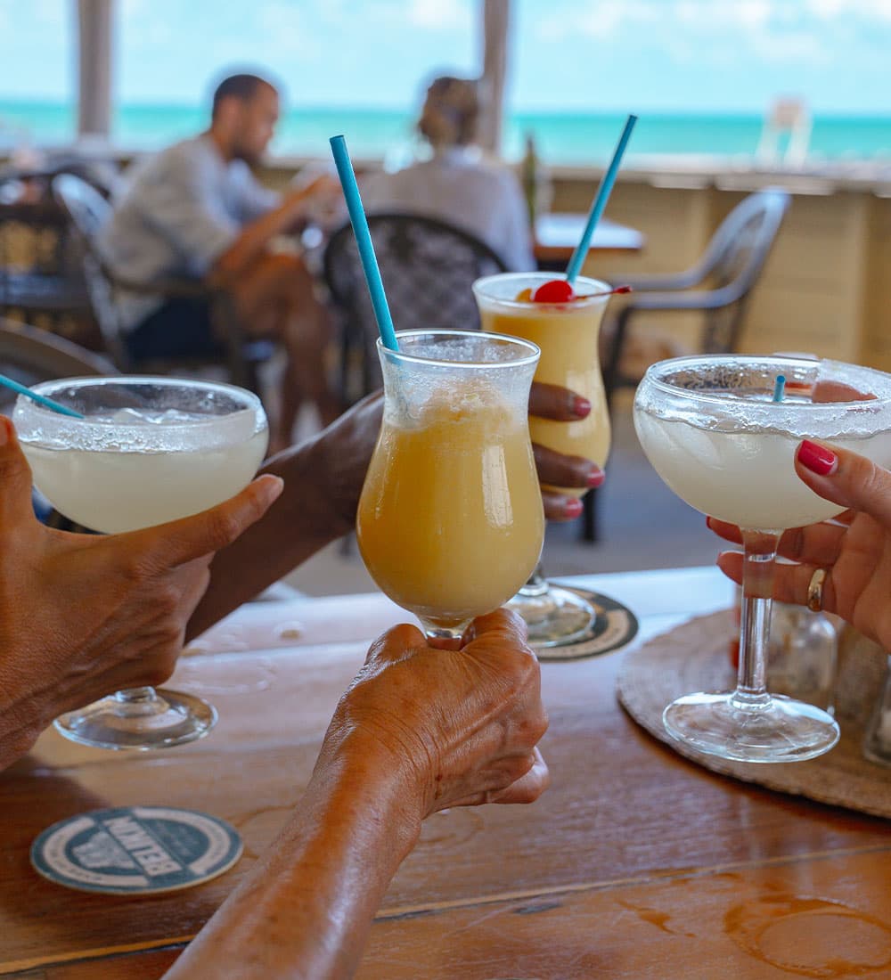 Three hands holding tropical cocktails with a beach backdrop.