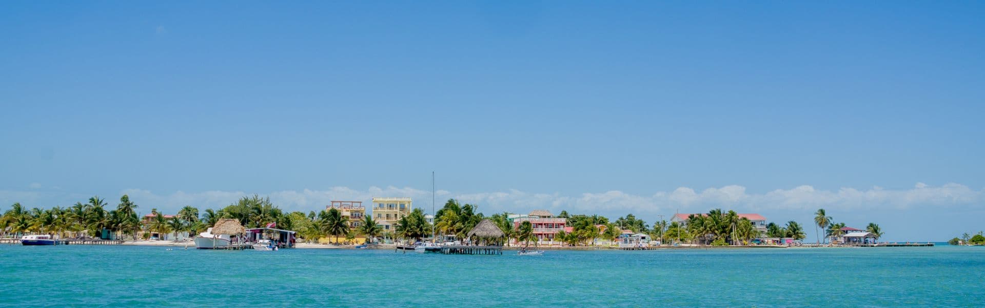 A tropical shoreline with palm trees and colorful buildings along a calm turquoise sea under clear blue skies.