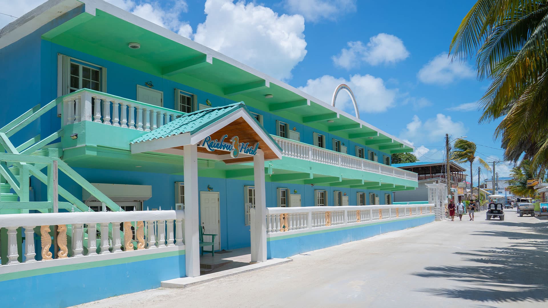 A colorful two-story hotel named "Rainbow Hotel" with a bright blue facade and palm trees lining the street.